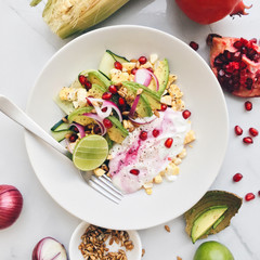 Plate of colorful healthy salad with avocado, sweet corn, pomegranate, cucumber, red onion, lime, greek yogurt and sunflower seeds on a marble background, top view