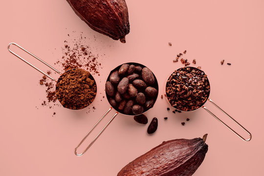 Rose Gold Measuring Cups Of Cocoa Beans, Cacao Nips, Cocoa Powder And Cocoa Pods On A Pink Background, Flat Lay Healthy Food Concept