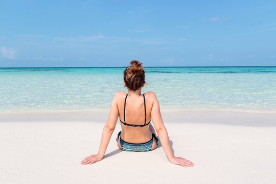 picture from back of a young woman seated on a white beach in the Maldives. Crystal clear blue water as background
