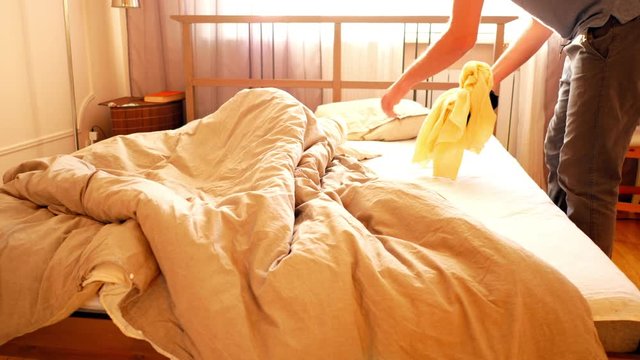 slow motion guy takes away scattered clothes of bed and makes double bed in bedroom lit with sunlight in morning