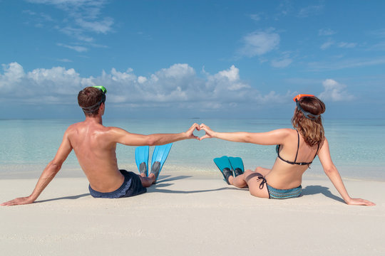picture from back of a young couple with flippers and mask seated on a white beach in the Maldives. Crystal clear blue water as background. Hand heart shape