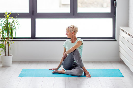 Mature woman in joga pose exercise in appartment at rug with window on background