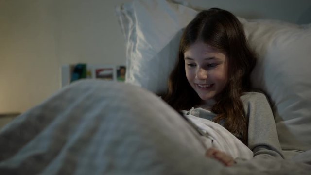 Modern, happy child sitting up in bed enjoying watching a movie on her electronic tablet.
