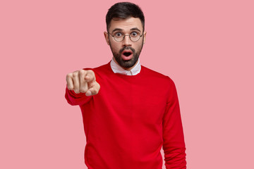 Terrific man with eyes popped out, points with index finger directly at camera, sees amazing scene, has bated breath, dark bristle, isolated over pink background. People, advertisement, reaction