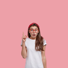 Vertical shot of amazed pretty teenager points with fore finger upwards, says look at this promo,...