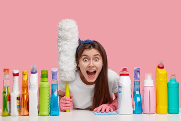 Emotive housemaid screams from panic, sees much work about house, going to clean dust on furniture with special brush, surrounded with cleansing products, stares at camera, isolated on pink wall