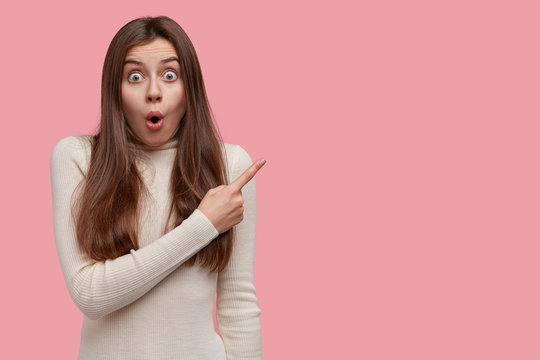 Beautiful young woman stares astonished, folds lips in wow sound, points right with index finger, demonstrates something surprising, isolated over pink background, shocked from incredible scene