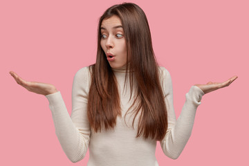 Photo of indecisive woman spreads hands, has clueless expression, doubts between two items, wears...
