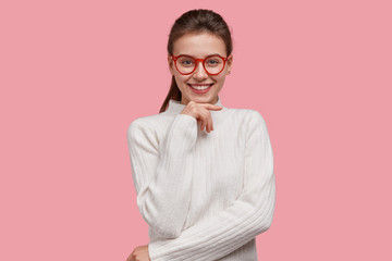 Positive young college student wears white winter sweater, red rim spectacles, keeps hand under chin, smiles broadly, being in good mood, isolated over pink background. Emotions and people concept