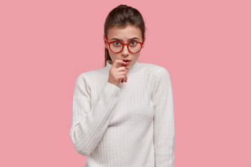 Isolated shot of serious pleasant looking woman keeps fore finger near lips, looks seriously through spectacles, dressed in white casual clothes, has doubtful gaze directly at camera, isolated on pink