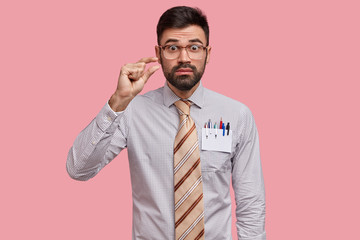 Photo of puzzled unshaven young man makes size gesture, demonstrates something tiny, wears formal shirt and long tie, has surpised expression, isolated over pink background. Omg, its too little