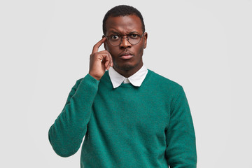 Puzzled dark skinned man frowns face, keeps index finger on temple, tries to concentrate, being deep in thoughts, wears green jumper, big spectacles, isolated over white background. Facial expressions