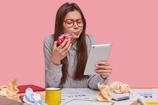 Image of serious beautiful lady holds delicious doughnut, watches training video on touchpad, prepares business report, studies graphic, drinks coffee, isolated over pink studio wall. Technology