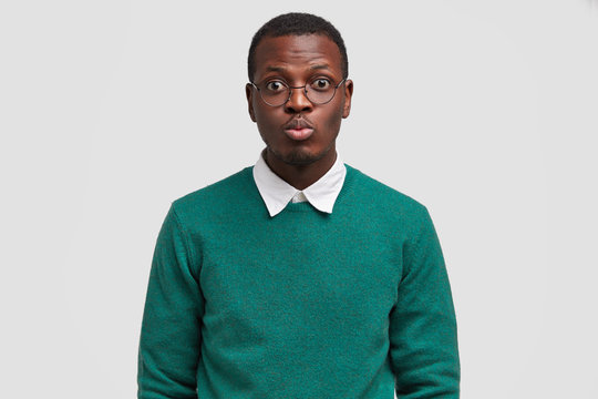 Serious young male hipster rounds lips for kiss, has date with woman, wears round glasses, dressed in green sweater, isolated over white background, has funny look, isolated on white background.