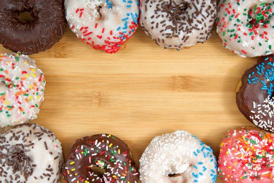 Flat lay view of many Frosted donuts with candy sprinkles arranged in a border frame on wood table with copy space.