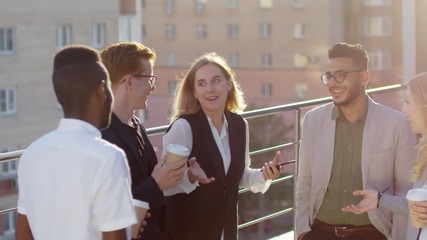 Cheerful blonde businesswoman holding smartphone and chatting with interracial young colleagues while standing on rooftop and having coffee break - Powered by Adobe