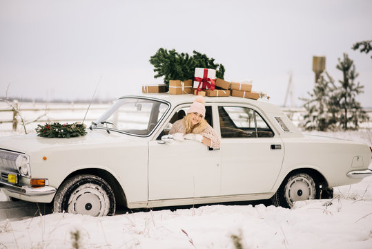 The girl rides in a retro car decorated with a Christmas tree and presents in a snowy forest. The concept of a winter Christmas photo shoot