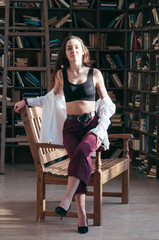 Attractive young female wearing black sport bra lingerie and white shirt unbuttoned, casual fashion style, sitting on wooden chair, bookshelves in the backdrop. Long hair, sensual beautiful seductive