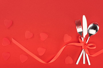 elevated view of cutlery wrapped by festive ribbon near heart symbols isolated on red, st valentine day concept