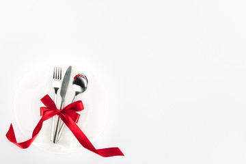 elevated view of fork, knife and spoon wrapped by red festive bow on plate isolated on white, st...