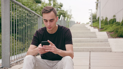 Fototapeta premium A young man using a phone sitting on the stairs in town. Medium shot. Soft focus