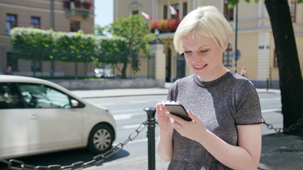 An attractive young blonde lady using a phone in town. Medium dolly shot. Soft focus