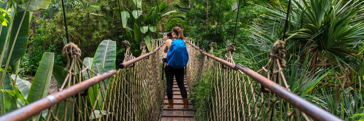 Woman with a backpack taking photographs on a suspension bridge in the jungle panorama