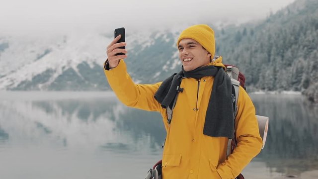 Handsome hiker man video calling on smartphone, waving at camera, smiling and speaking with friends. Beautiful winter mountains and lake against background, Slow motion