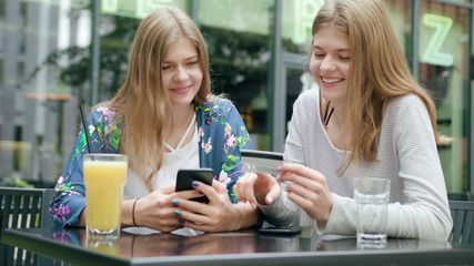 Attractive young ladies sitting at the table outdoors and using a phone and holding a credit card. Medium shot. Soft focus