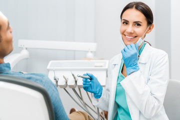 cheerful female dentist holding drill and smiling near patient - Powered by Adobe