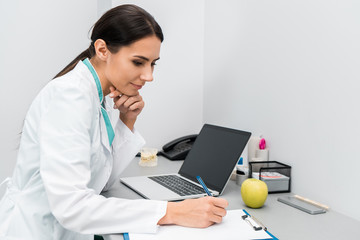 thoughtful female doctor making notes near laptop