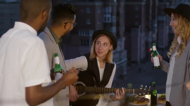 Young hipster woman playing the guitar and singing a song to her multi-ethnic friends standing around with beer bottles and enjoying the music at summer party on urban rooftop in the night