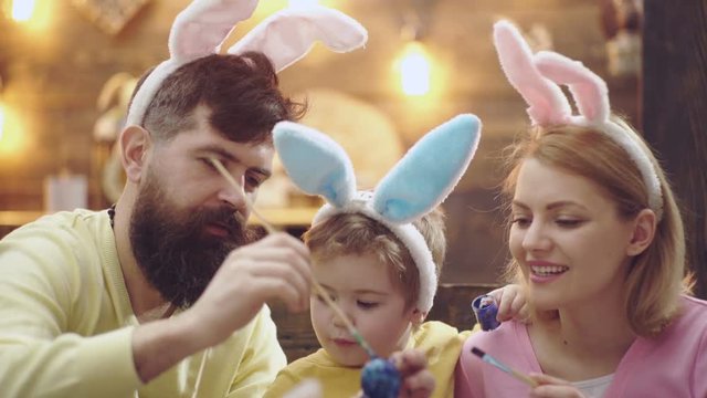 Happy family are preparing for Easter. Cute little boy wearing bunny ears. Concept of Happy Easter. Bearded man with a blond woman and a little boy dressed in a hares ears are painting Easter eggs.
