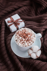 Coffee cup with marshmallow and gift box with brown ribbon on warm knitted brown blanket.
