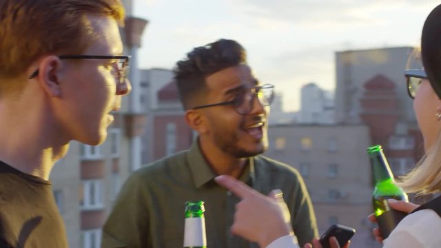 Young multiethnic friends dancing with beer bottles and smiling while having party on urban rooftop at sunset; middle eastern man using smartphone