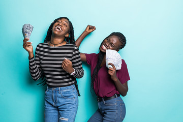Portrait of two african delighted girls rejoicing win and dancing while holding fans of cash money...