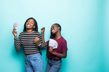 Two pretty african young girls friends standing showing money banknotes, celebrating isolated over green background