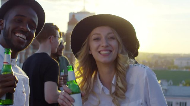 Young african american man and beautiful caucasian woman in fedora hats holding beer bottles, smiling and looking at camera while dancing together at rooftop party with friends