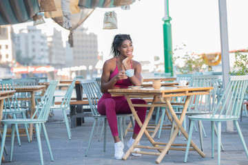 Beautiful tall athletic African American Woman wearing a bright pink workout outfit sits in an outdoor cafe drinking coffee or tea in deep though on a bright sunny day