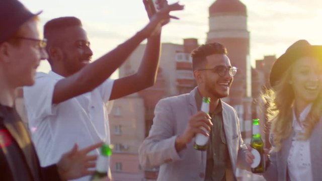 Group of interracial young men and women smiling and dancing with beer bottles while having outdoor party on urban rooftop at sunset