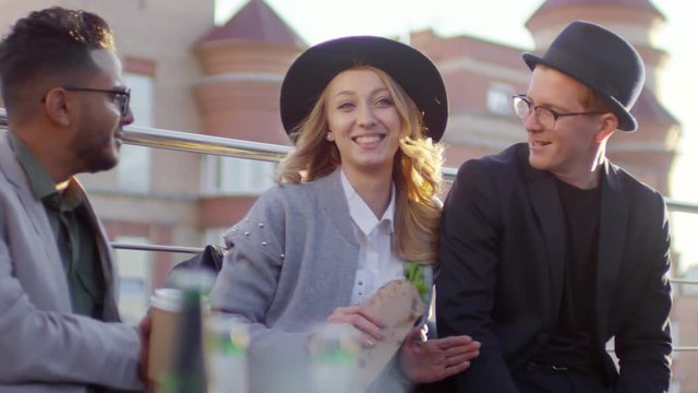 Young beautiful woman in fedora hat holding sandwich, smiling and chatting with male multiethnic friends while having lunch on rooftop