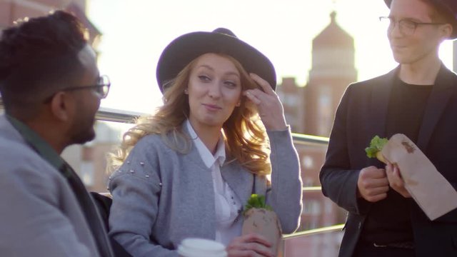 Young beautiful woman in fedora hat holding sandwich and speaking with male friends while having lunch on rooftop; sunlight shining at camera