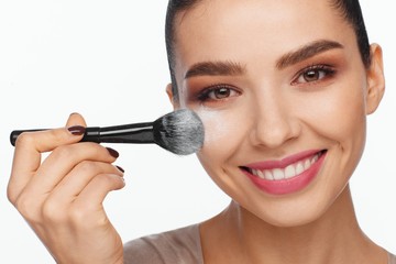 Portrait of a beautiful smiling young woman holding a brush in her hand and applying powder to her face with a brush
