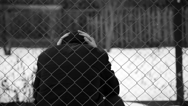 Young unidentifiable teenage boy holding hes head at the correctional institute in black and white, conceptual footage of juvenile delinquency, focus on the wired fence in black and white.