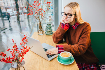Cozy workplace in rainy day - teenager student woman in eyeglasses wearing red sweater with laptop drinking coffee in a cafe alone near big window in modern interior small coffee shop during work time