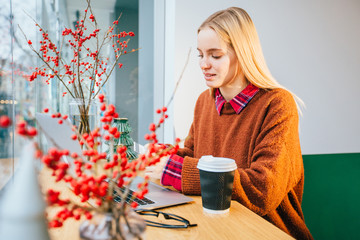 Portrait of beautiful blond hipster freelancer business woman in terracotta sweater shopping online while sitting in a cafe with laptop. Receive payment concept.