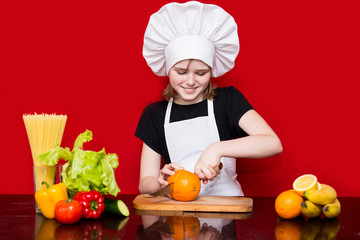 Happy little girl in chef uniform cuts fruit in kitchen. Kid chef. Cooking Concept