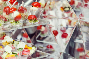 Catering for party. Close up of appetizers with cherry tomatoes, green olives, olive oil, cheese and spices in short glasses on wood brown table. Horizontal color image.