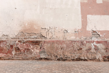 Old dirty damaged wall with cracks, chips, peeling white and pink paint. Brick wall and part of floor in terrible slums. Grunge Red Stonewall Background. Shabby Building Facade With Damaged Plaster.