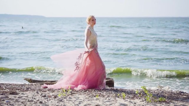 attractive lady on beach enjoys the wind, holds light tulle plume with her fingers, dreams, listens to the sound of the ocean, waves, blonde beauty in long luxurious pink dress, fabulous image 0016 